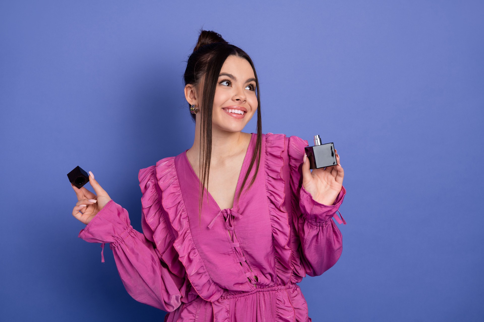 Charming young woman in pink ruffled dress with perfume bottle on trendy purple backdrop